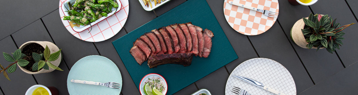 Steak on a blue cutting board with side dishes and cutlery on a wooden table