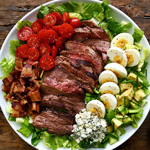 Plated salad with steak, tomatoes, eggs, bacon, and avocado on a wooden surface