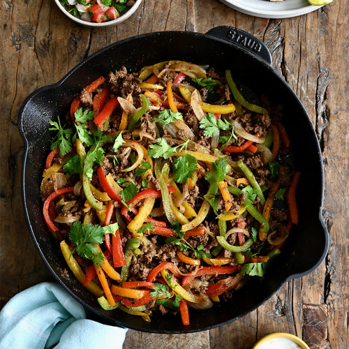 Skillet with ground beef, bell peppers, and cilantro on a wooden table.
