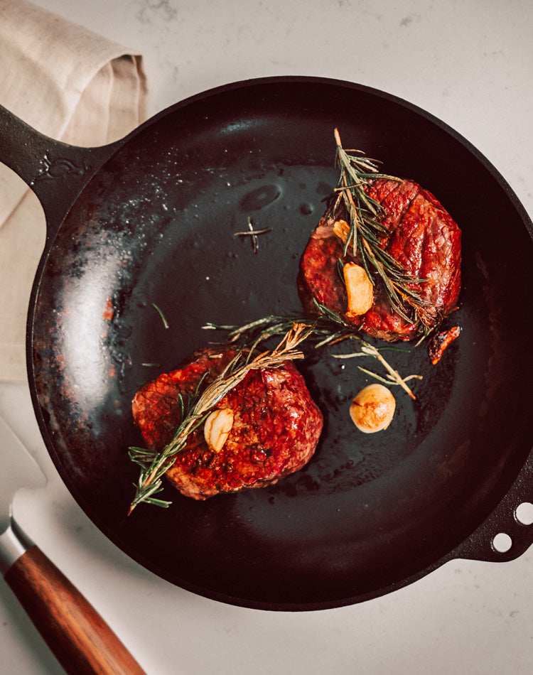 Two steaks with rosemary and garlic in a cast iron skillet on a light background