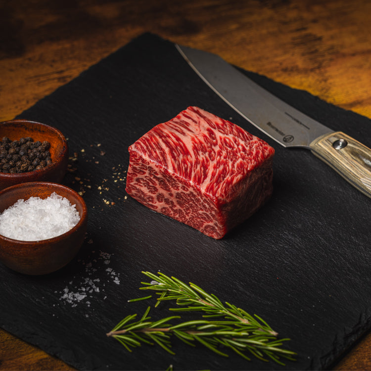 Raw steak on a black slate board with a knife, salt, pepper, and rosemary.