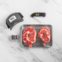 Two raw steaks on a grill pan with a cap, container, and thermometer on a marble surface.