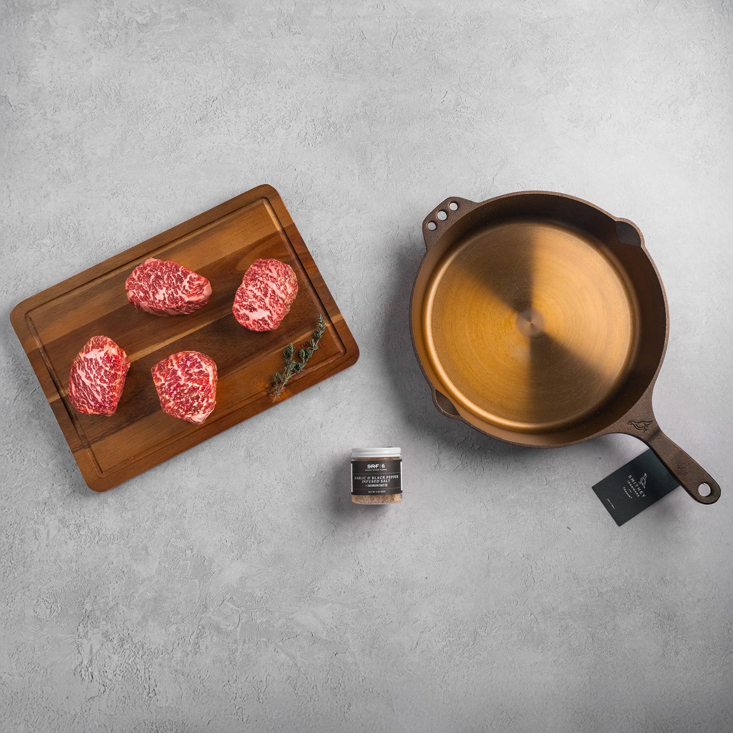 Skillet, wooden cutting board with raw meat, and seasoning on a gray surface