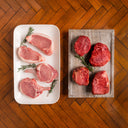 Raw pork chops on a white plate and raw steak on a wooden board with a wooden floor background.