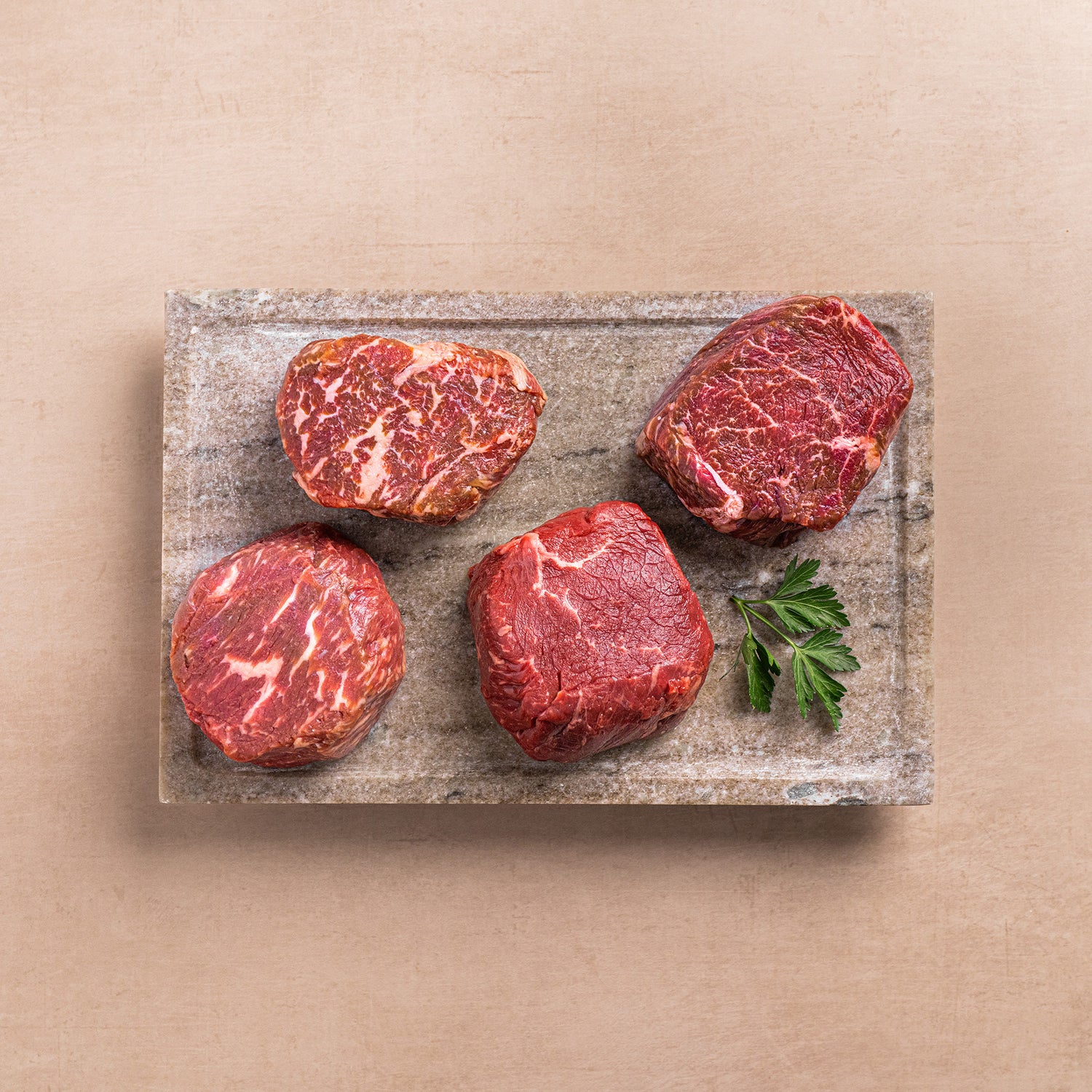 Four raw beef steaks on a stone cutting board with a leaf on a beige background