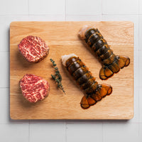 two wagyu ribeye filet steaks and two lobster tails on a cutting board on a white tile background