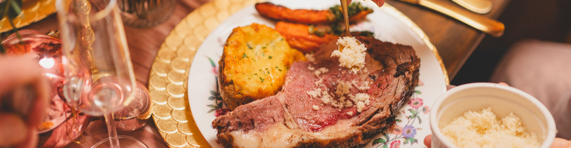 Plate of sliced prime rib with carrots, potatoes, and a side of mashed potatoes on a floral tablecloth.