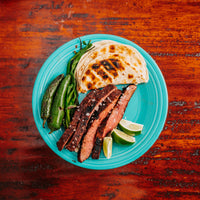 cooked sliced flank steak on plate with tortilla shells, peppers, and limes