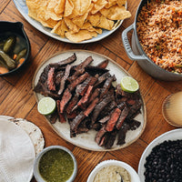 cooked outside skirt steak on table with chips, rice, beans and other sides 