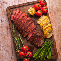 cooked and sliced flat iron steak on cutting board with veggies and bread 