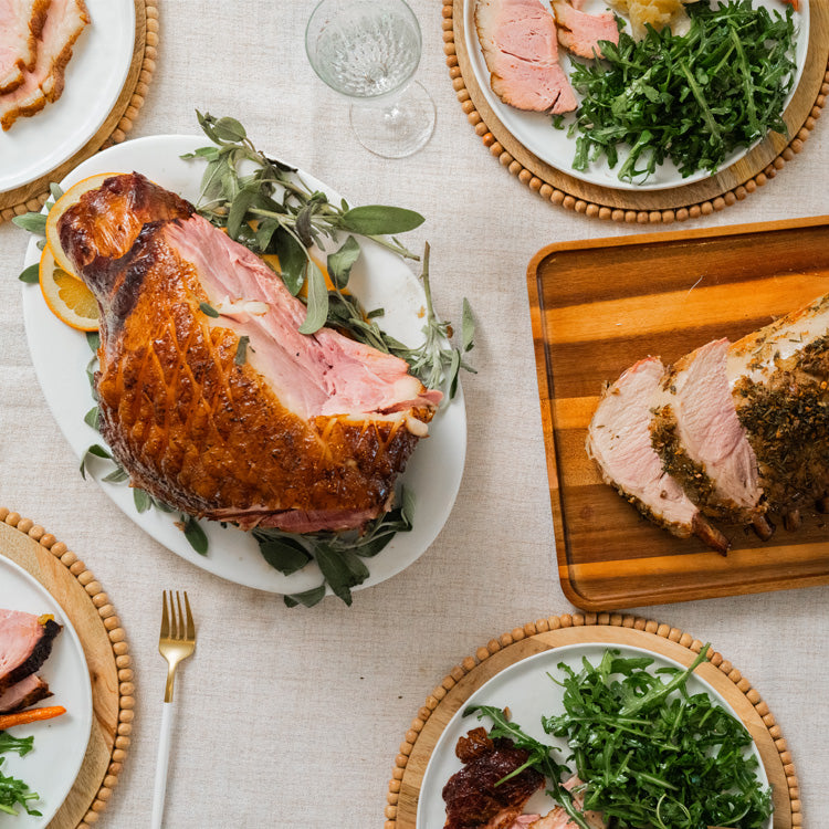 Roasted Kurobuta ham with greenery on a plate, surrounded by other dishes and cutlery on a light surface.