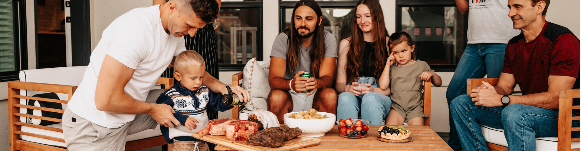 family around an outdoor table seasoning steaks with other dishes 