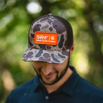Person wearing a camouflage cap with an orange SRF logo against a blurred green background