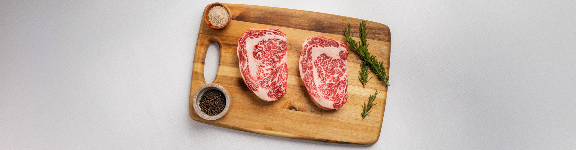 Two raw ribeye steaks on a wooden cutting board with spices and herbs.