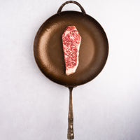 Raw Gold Grade NY steak on a cast iron skillet with a white background