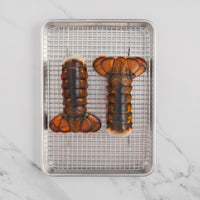 Two lobster tails on metal sheet pan with grate.