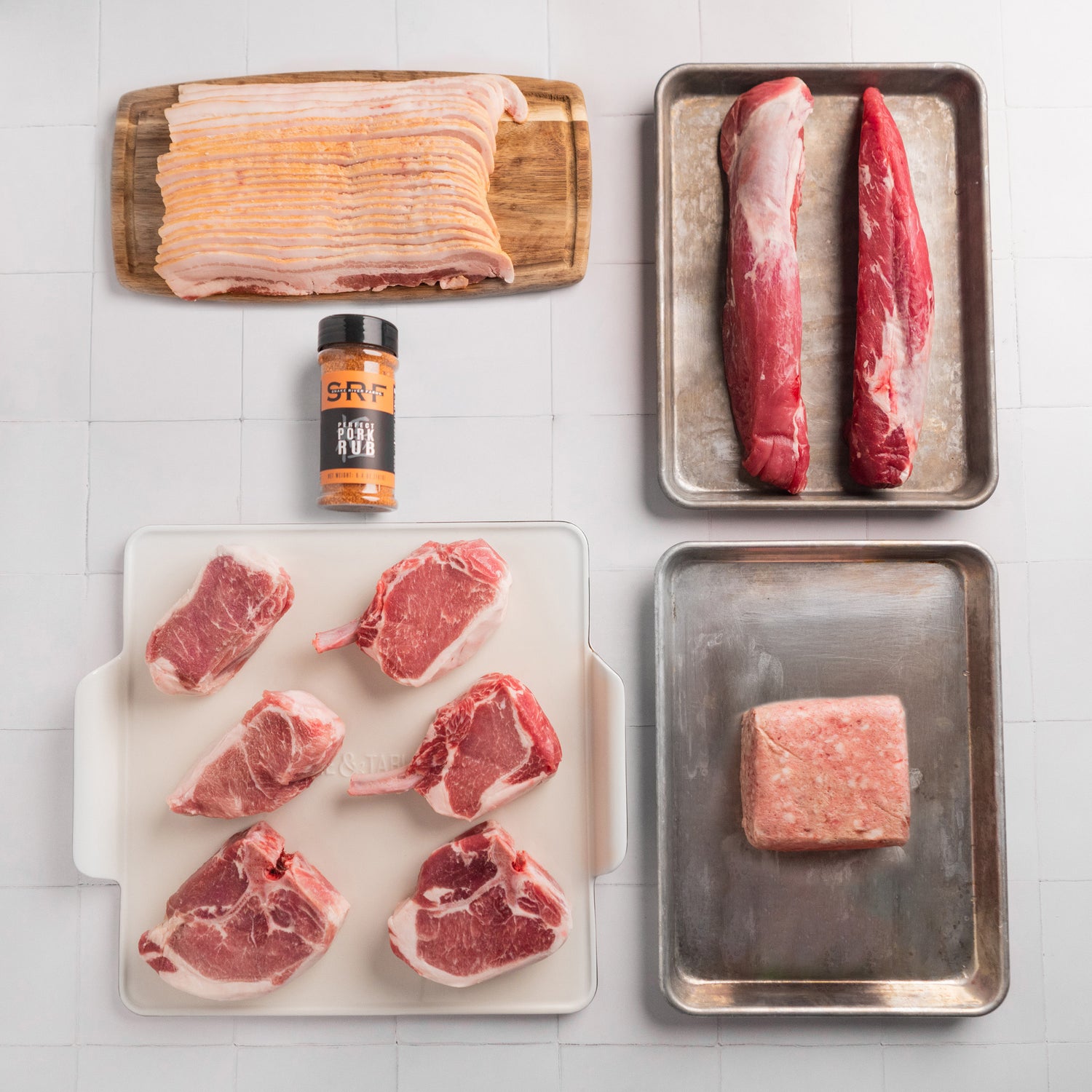Assorted raw meats on trays with a seasoning container on a tiled surface