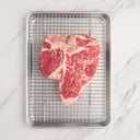A dry-aged porterhouse steaks on a wire rack inside a metal baking tray, on a marble countertop.