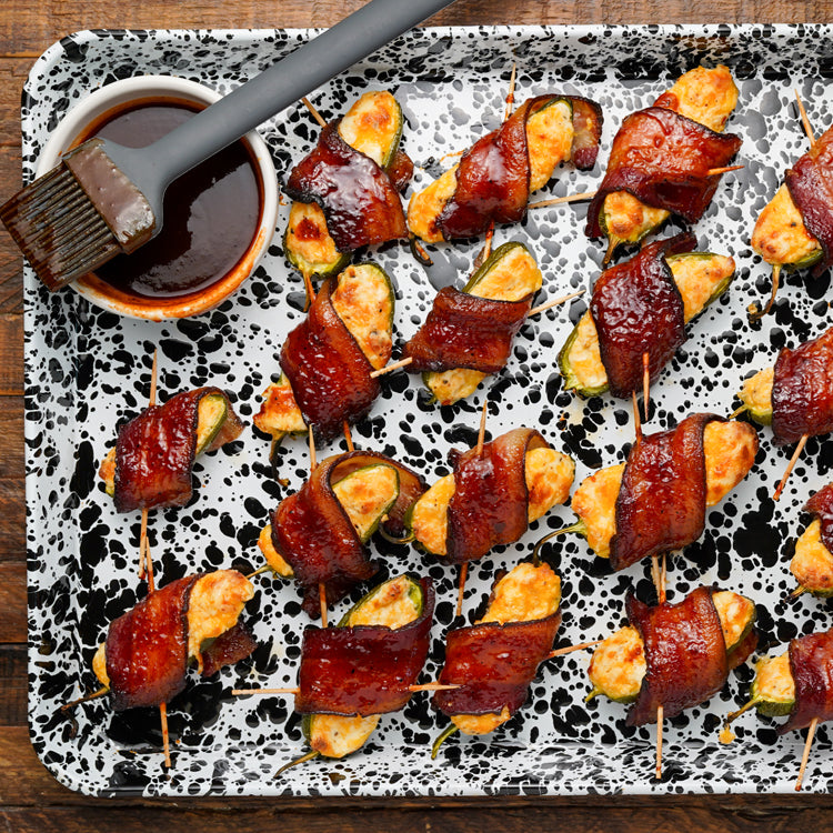 Bacon-wrapped vegetable skewers on a speckled tray with a brush and sauce.