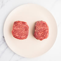 Two raw steaks on a white plate with a marble background