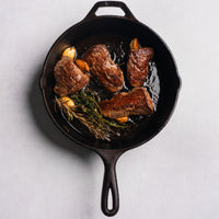 Sear marks on beef pieces in a cast iron skillet with herbs and garlic on a light gray background