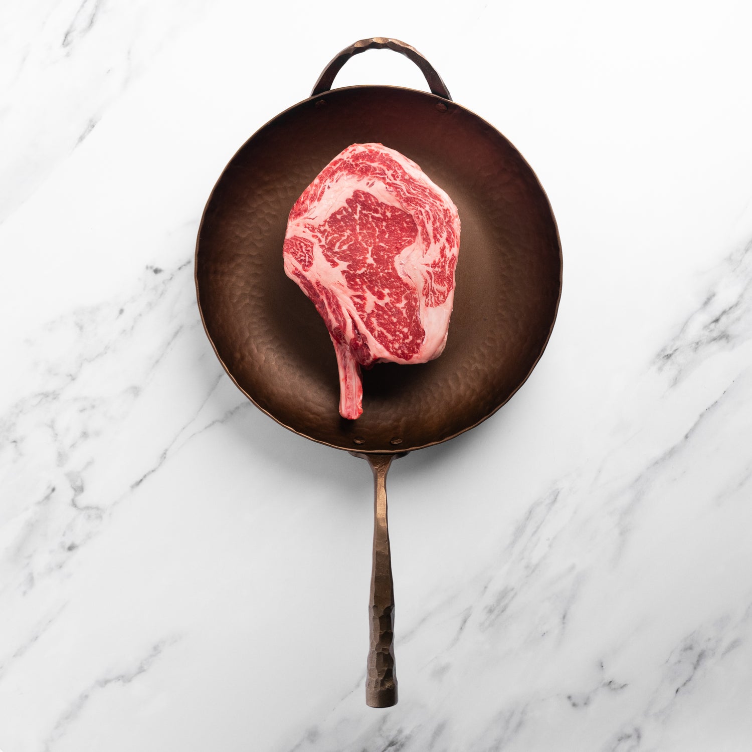 Raw cowboy ribeye steak in a frying pan on a marble surface