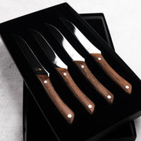 Set of four steak knives with wooden handles in a black box on a light gray surface.
