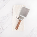 A stainless steel spatula with a full tang walnut handle, displayed on a beige cloth with the Smithey logo.