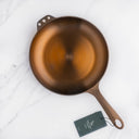 A top view of a bronze-colored cast iron skillet with a chef-approved handle, displayed on a marble surface with a visible product tag.
