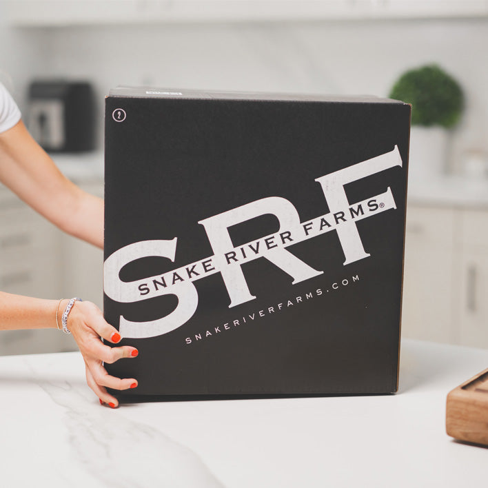 Person holding a black box with 'Snake River Farms' branding in a kitchen setting