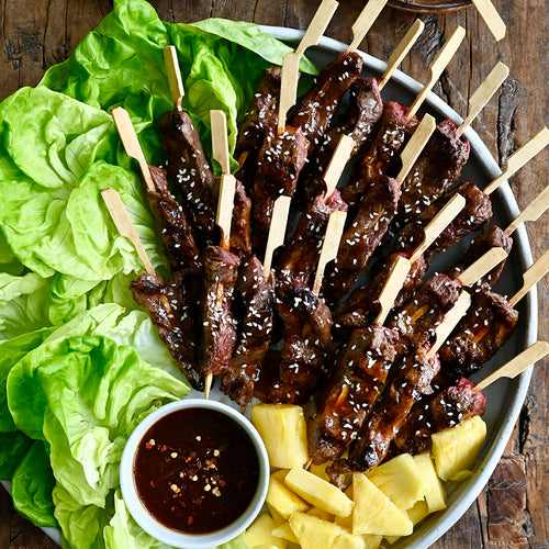 Skewers of marinated steak with pineapple chunks on a platter with lettuce and dipping sauce.