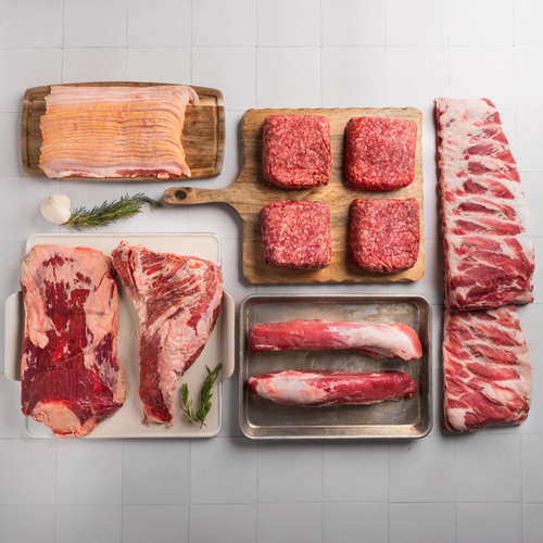 Assorted raw meats including bacon, steaks, ribs, and a ribeye on a tiled surface.