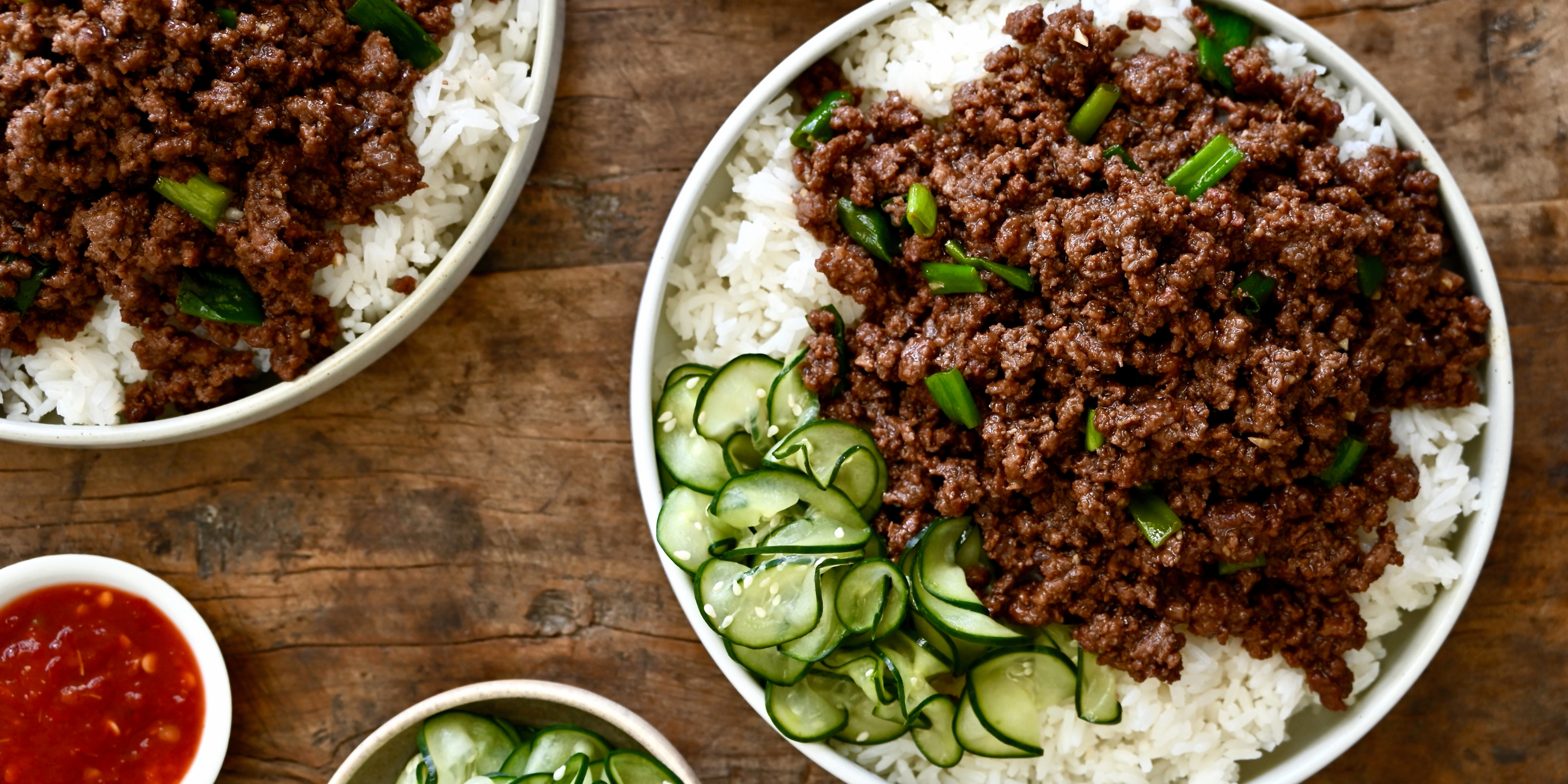 Overhead view of a bowl of white rice topped with brown ground meat and green onions, with sliced cucumber on top, served on a rustic wooden table. A small side dish of red sauce is visible at the lower left.