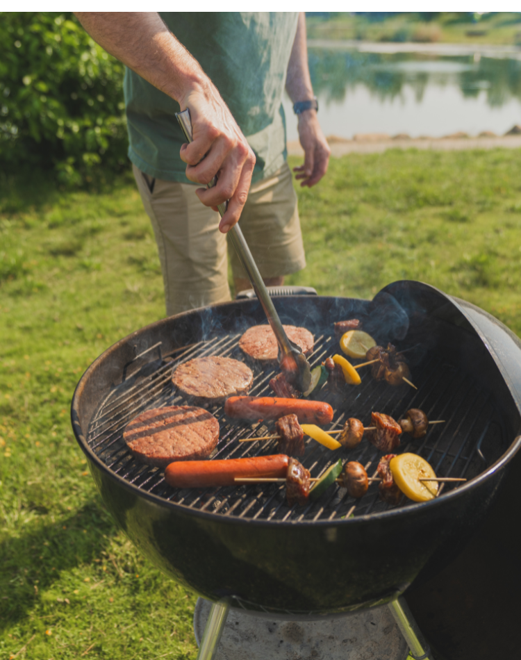 A person grilling outdoors on a black round charcoal grill, with burger patties, sausages, and skewers cooking over the flames. The scene is set on a grassy lawn with a lake in the background.