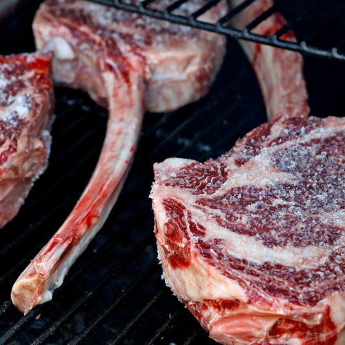 Raw tomahawk steaks with visible marbling and coarse salt crystals, resting on dark grill grates.