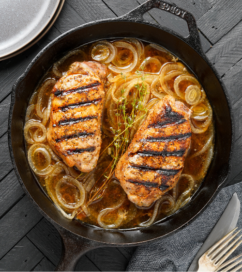Overhead view of a cast iron skillet containing two grilled Kurobuta pork chops with visible grill marks, cooked in a brown sauce with caramelized onions and a sprig of thyme, set on a dark wooden surface.