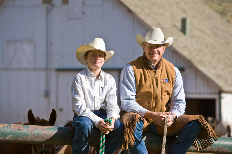 Man and young boy sitting on a fence with horses in the background