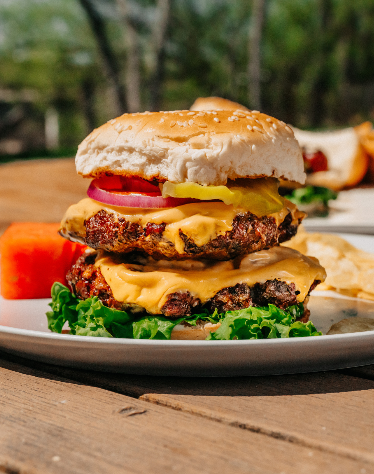 Double cheeseburger with lettuce, tomato, and onion on a wooden table with blurred greenery in the background.