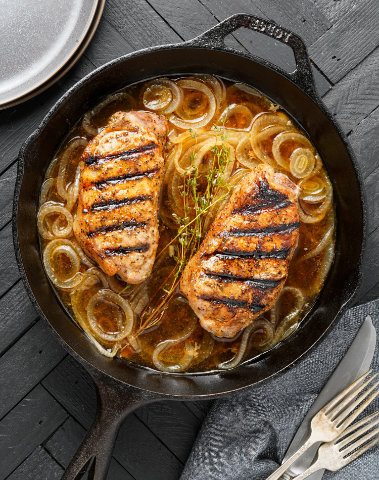 Two grilled pork chops in a skillet with onions on a dark wooden surface.