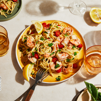 Platter of shrimp pasta with lemon wedges on a table with drinks and garnishes.