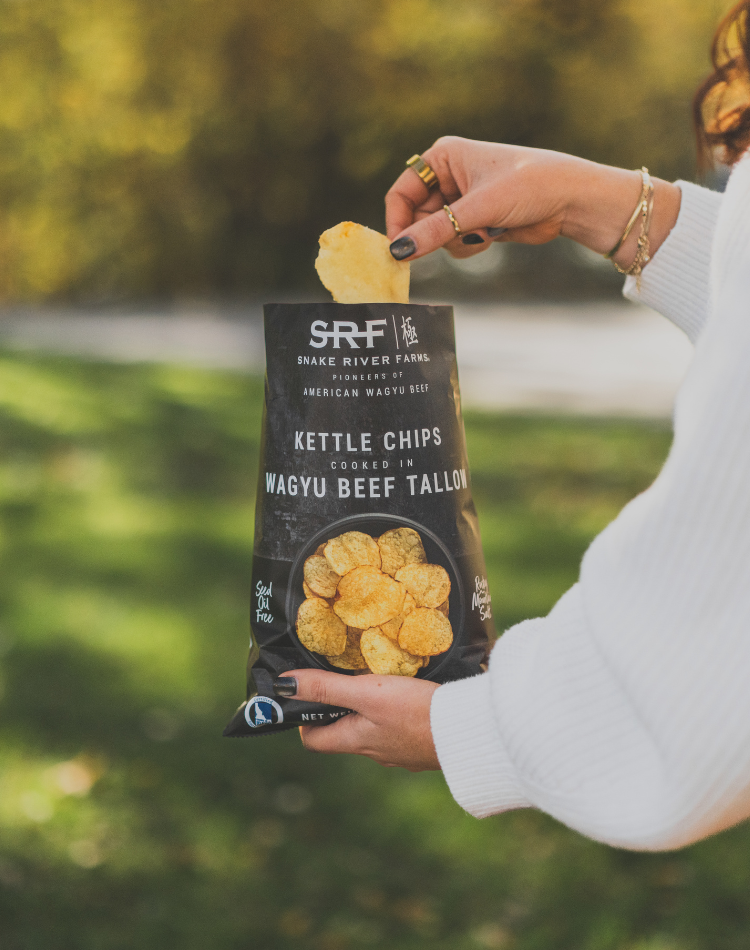 Person holding a bag of Snake River Farms kettle chips with a blurred outdoor background