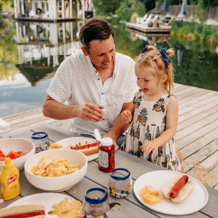 Man and young girl at a lakeside picnic with food and drinks.