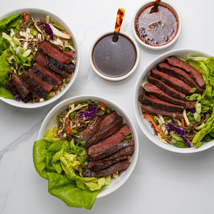 Three bowls of steak salad with side dishes on a marble surface