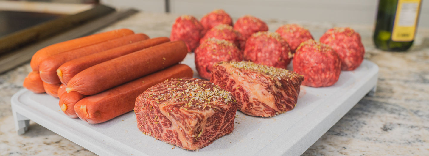 Assorted meats including hot dogs and beef on a white cutting board with a kitchen background.