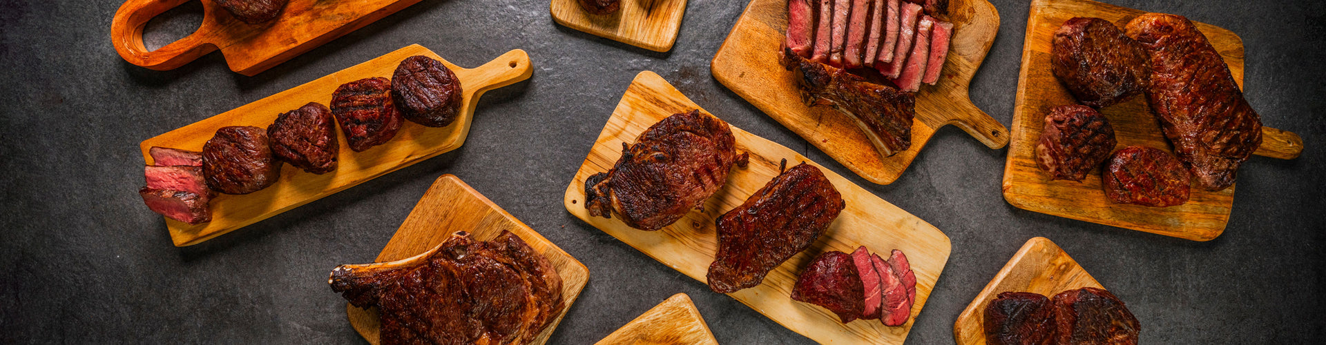Assorted cooked steaks on wooden cutting boards against a dark background