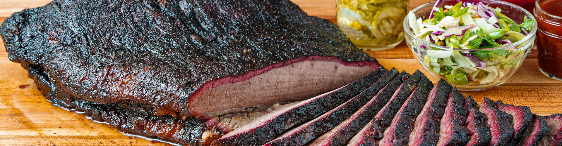 Smoked brisket on a wooden cutting board with sides of pickles and coleslaw.
