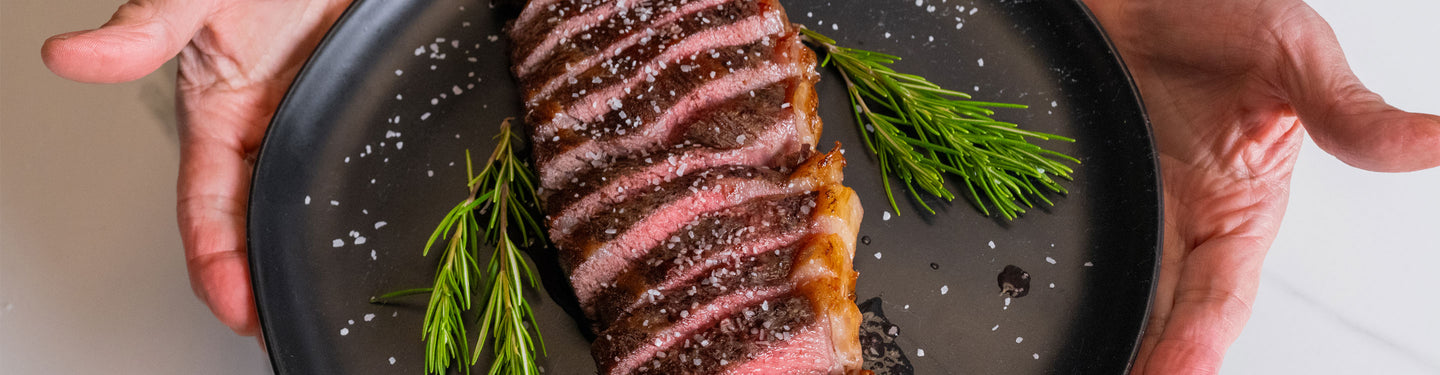 Steak on a plate with rosemary, held by two hands against a neutral background