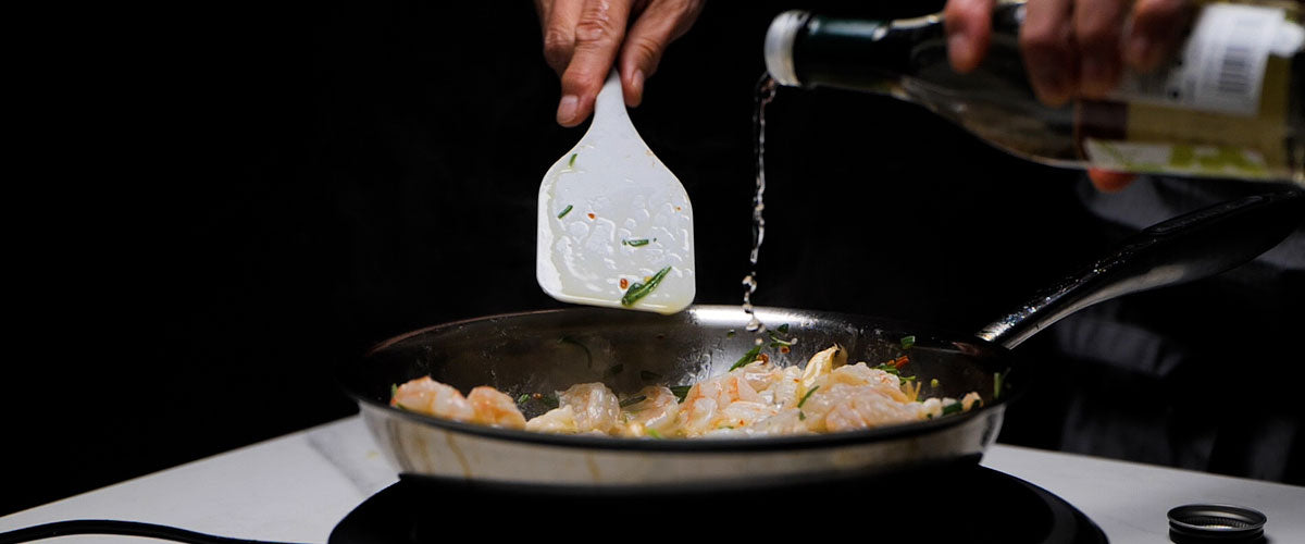 Person cooking shrimp in a pan with a spatula and oil bottle.