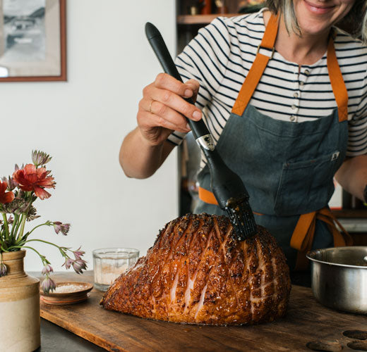 Person in an apron preparing a roasted ham on a wooden cutting board.
