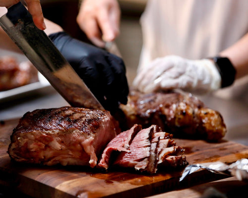 A close-up of a person slicing a cooked steak on a wooden cutting board, wearing black and white gloves while using a large knife.